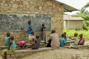 Children learning in an outdoor classroom setting with a chalkboard and stone wall backdrop.